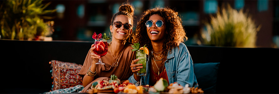 Duas jovens mulheres felizes tomando drinks coloridos em um ambiente ao ar livre durante o dia, usando óculos de sol e roupas casuais, desfrutando de uma festa ou encontro social.
