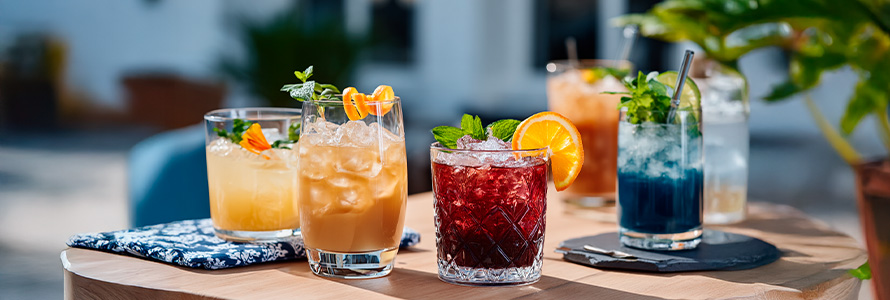 Variedade de drinks coloridos e refrescantes em copos de vidro, decorados com frutas e hortelã, em um ambiente ao ar livre, ideal para happy hour.