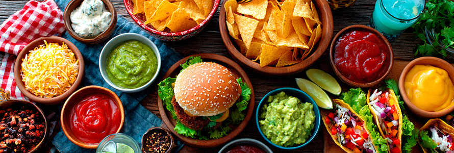 Variety of Mexican foods including mini burgers, tacos, guacamole, nachos and different colorful sauces on wooden table