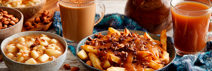 A plate of fries topped with gravy, served with two glasses of carrot juice and a hot drink, with additional ingredients on the side.
