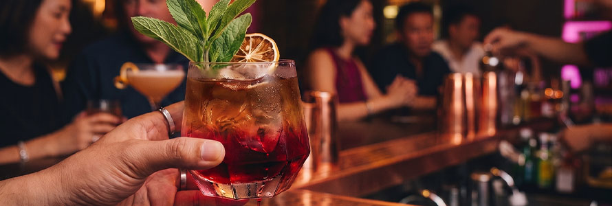 Mano sosteniendo cóctel rojo con menta en barra de bar iluminada